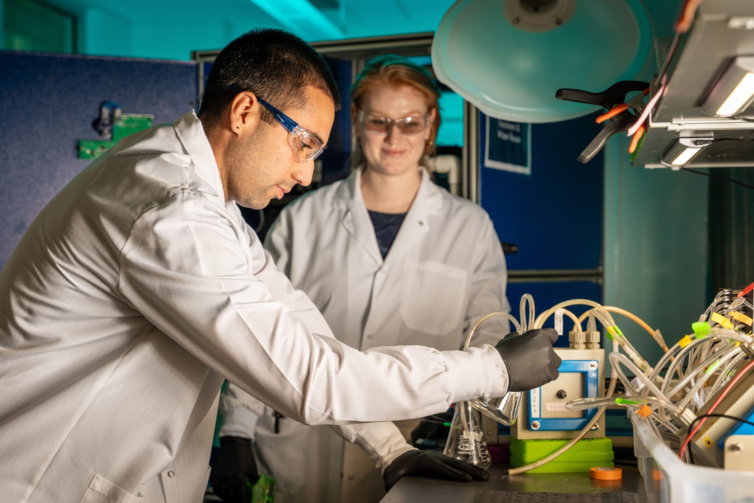 A man and a woman in a laboratory wearing white coats and safety goggles, putting something in a flask.