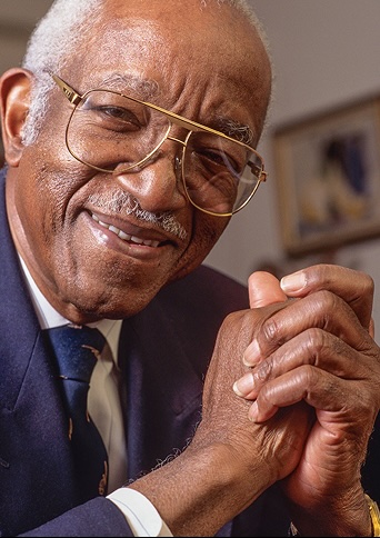 An older gentleman with wide glasses and white receding hair in a blue suit and tie smiles at the camera while grasping his hands together.