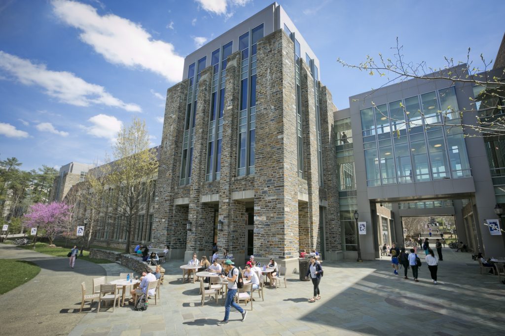 College students walking around and eating at cafe tables in front of a campus building on a sunny day.