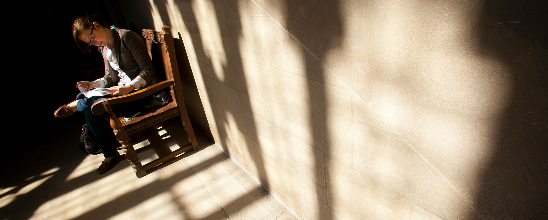 A student reading a book sits on an ornate bench next to a sandstone wall.