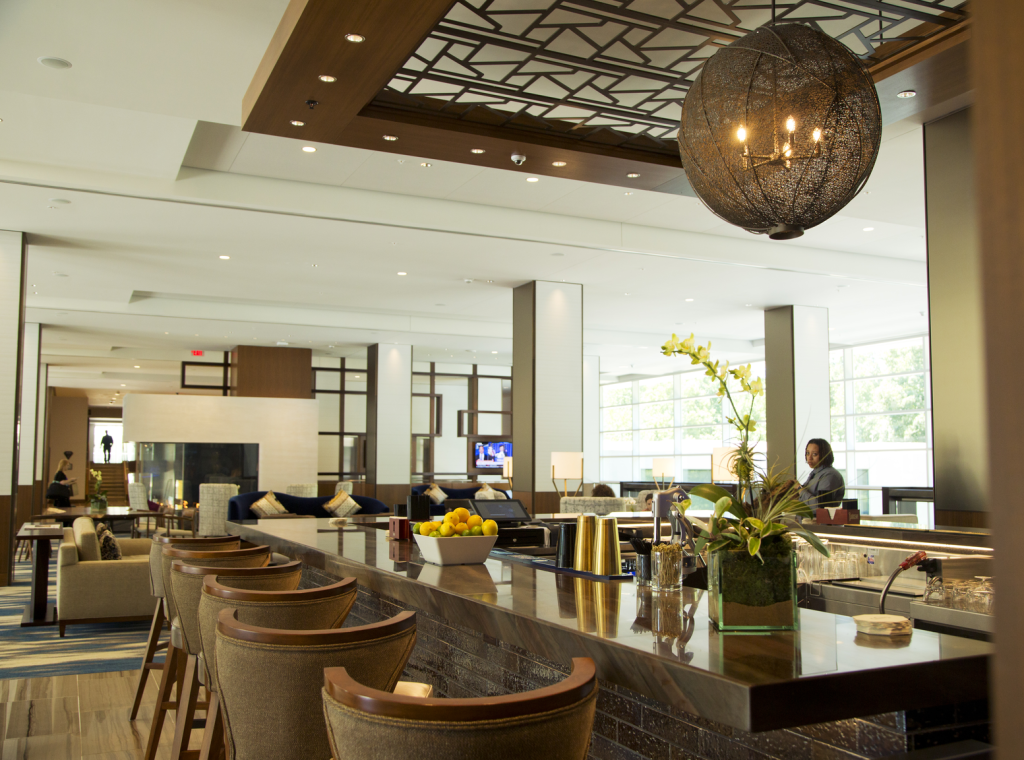 Upscale lobby bar with stools, a large metallic chandelier and decor on the bar surface, like a bowl of lemons and limes, and orchids. A fireplace and couches in the background.