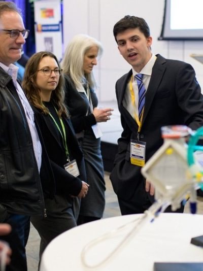 Four people in suits at an innovation fair looking at a table with a device made of tubes.