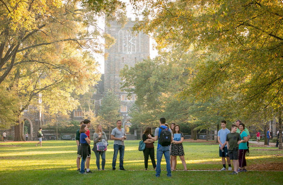 About a dozen young people on a campus tour listen to a tour guide on a sunny day on a green campus quad.