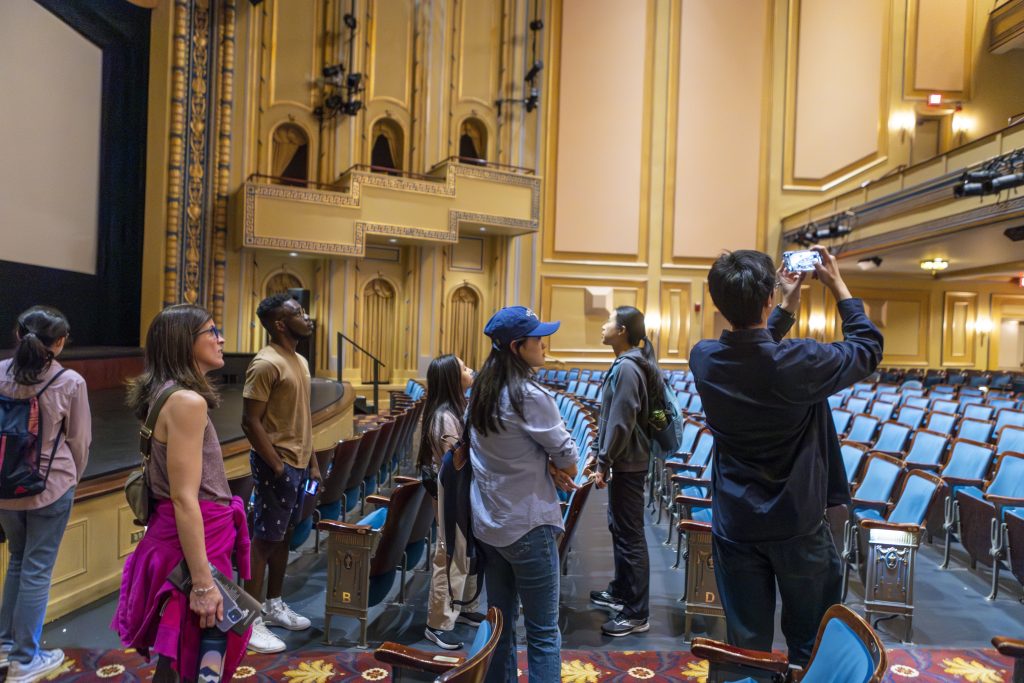 Six students in an empty Carolina Theater, standing around the folding seats, one taking a photo with his iPhone.