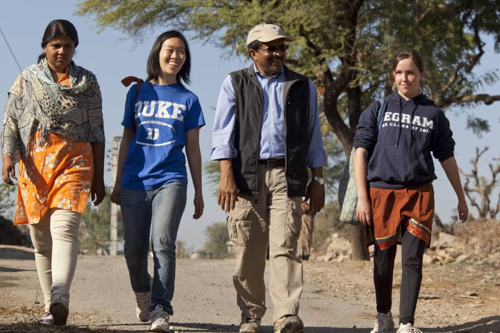 Four students walking down a dirt road with a tree in the background.
