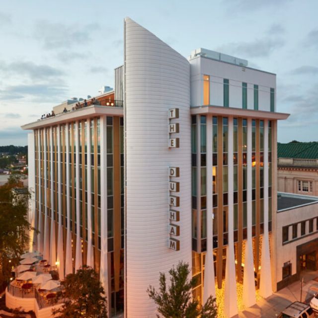 Aerial view of an art deco style hotel with lots of windows and vertical columns and a warmly lit lobby inside. Letters vertically running down the building spell "The Durham."