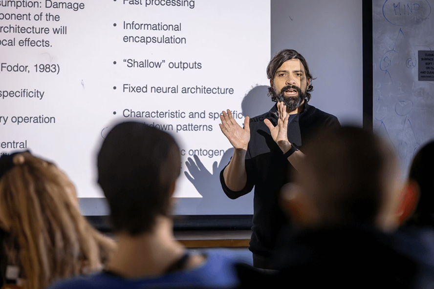 A male professor with dark hair and beard in a black collared shirt raises his hands to explain a concept in front of a screen with a presentation on it.