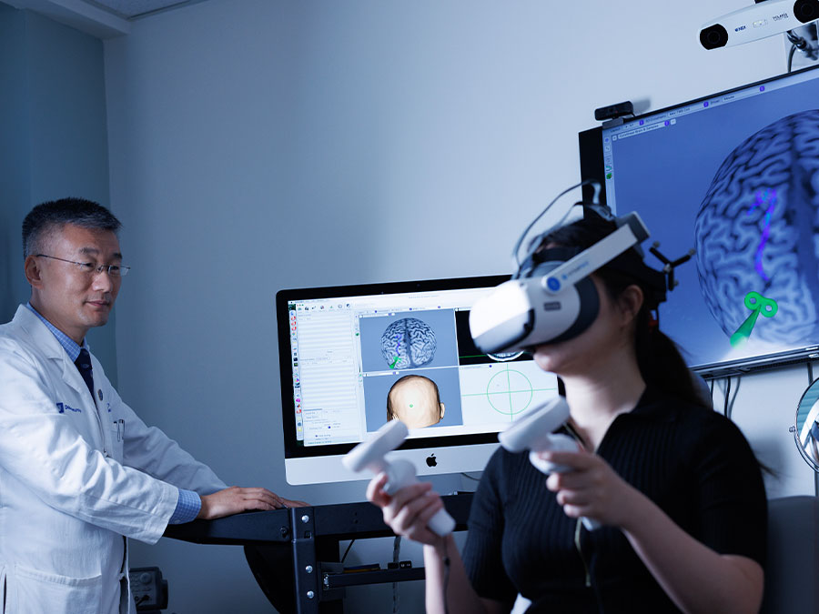 A doctor looks on as a person with a virtual reality headset on sits in a chair with images of brains on the screens in the background